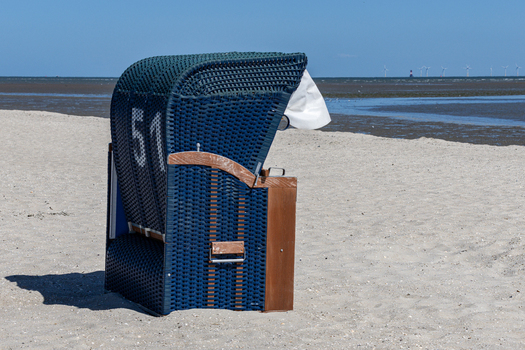 View of a blue wicker beach chair numbered 51 on the sandy shore of Strand Schillig with the Wadden Sea and wind turbines in the background Wangerland Niedersachsen Germany. View of a blue wicker beach chair numbered 51 on the sandy shore of Strand Schillig with the Wadden Sea and wind turbines in the background Wangerland Niedersachsen Germany.