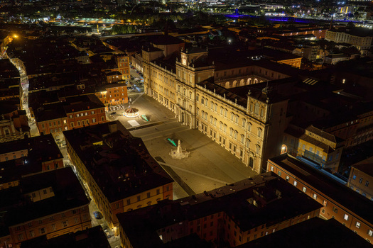 Aerial view of the illuminated Palazzo Ducale di Modena in Piazza Roma at night surrounded by historic city buildings and street lights in Modena Emilia Romagna Italy.