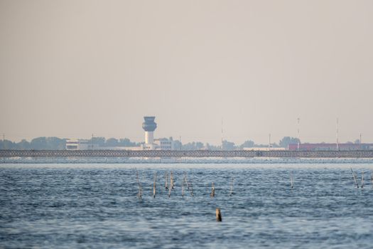 View of the calm waters of Narta Lagoon with a distant control tower and shoreline buildings under a hazy sky in Zvernec Vlore County Albania.