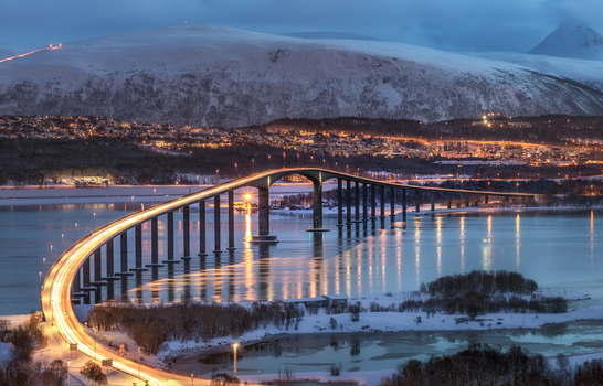 View of the illuminated Tromso Bridge spanning the fjord with snow covered mountains and city lights under a twilight sky in Tromso Troms Norway. View of the illuminated Tromso Bridge spanning the fjord with snow covered mountains and city lights under a twilight sky in Tromso Troms Norway.