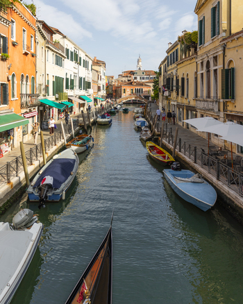 View of a narrow canal lined with historic buildings moored boats and a gondola in the foreground under a bright blue sky in Venice Veneto Italy. View of a narrow canal lined with historic buildings moored boats and a gondola in the foreground under a bright blue sky in Venice Veneto Italy.