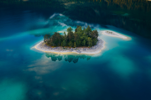 Aerial view of a small forested island surrounded by turquoise water and white rocky shores at Eibsee lake in Grainau Bavaria Germany. Aerial view of a small forested island surrounded by turquoise water and white rocky shores at Eibsee lake in Grainau Bavaria Germany.
