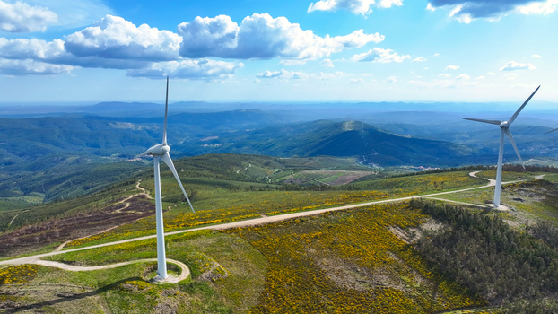 Aerial view of wind turbines standing on a rolling green hillside with yellow wildflowers and dirt paths under a bright blue sky in Lisbon Lisbon Portugal. Aerial view of wind turbines standing on a rolling green hillside with yellow wildflowers and dirt paths under a bright blue sky in Lisbon Lisbon Portugal.