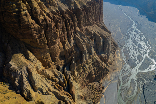 Aerial view of a massive rugged mountain cliff face towering over a wide braided riverbed in the arid landscape of Nepal.