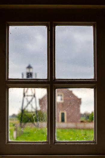 View of the Schokland lighthouse and a historic brick building seen through a dusty four pane window frame under a cloudy sky in Schokland Flevoland Netherlands.