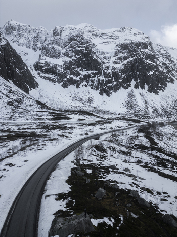 Aerial view of the winding road through the snowy landscape of Grotfjord with steep rocky mountains under a cloudy sky in Tromvik Troms Norway. Aerial view of the winding road through the snowy landscape of Grotfjord with steep rocky mountains under a cloudy sky in Tromvik Troms Norway.