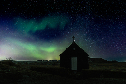 View of the black church silhouetted against the vibrant green and purple aurora borealis lighting up the starry night sky Krysuvikurkirkja Iceland.
