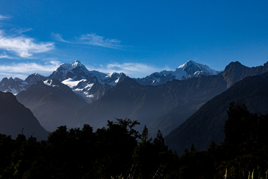 View of majestic snow capped mountains pierce the vibrant blue sky framed by the dark silhouettes of trees in the foreground Lake Matheson New Zealand. View of majestic snow capped mountains pierce the vibrant blue sky framed by the dark silhouettes of trees in the foreground Lake Matheson New Zealand.