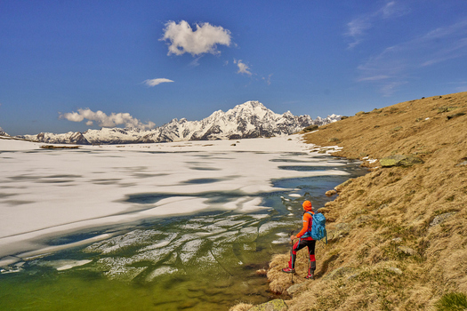 View of a hiker in orange gear standing by a partially frozen alpine lake with snow capped mountains in the background in Valmalenco Lombardia Italy. View of a hiker in orange gear standing by a partially frozen alpine lake with snow capped mountains in the background in Valmalenco Lombardia Italy.