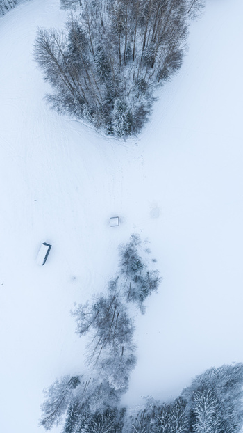 Aerial view of a snow covered landscape in Latvia featuring scattered trees and small structures under a winter sky. Aerial view of a snow covered landscape in Latvia featuring scattered trees and small structures under a winter sky.