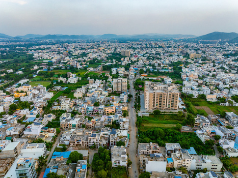 Aerial view of the cityscape of Udaipur Rajasthan India featuring residential buildings green spaces and a central road stretching towards the horizon. Udaipur Rajasthan India. Aerial view of the cityscape of Udaipur Rajasthan India featuring residential buildings green spaces and a central road stretching towards the horizon. Udaipur Rajasthan India.