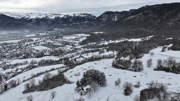 Aerial view of a small chapel nestled among snow covered trees and fields with the village and Jura mountains in the background Vesancy Auvergne Rhone Alpes France. Aerial view of a small chapel nestled among snow covered trees and fields with the village and Jura mountains in the background Vesancy Auvergne Rhone Alpes France.