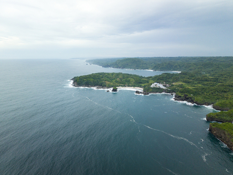 Aerial view of the rugged coastline where deep blue waters meet verdant cliffs and a hidden sandy cove under a dramatic sky Gunungkidul Regency Special Region of Yogyakarta Indonesia. Aerial view of the rugged coastline where deep blue waters meet verdant cliffs and a hidden sandy cove under a dramatic sky Gunungkidul Regency Special Region of Yogyakarta Indonesia.