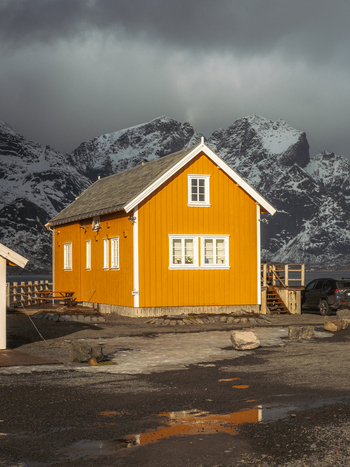 View of a vibrant yellow wooden house stands in stark contrast against the rugged snow dusted mountains under a brooding sky Hamn&oslash;y Nordland Norway.