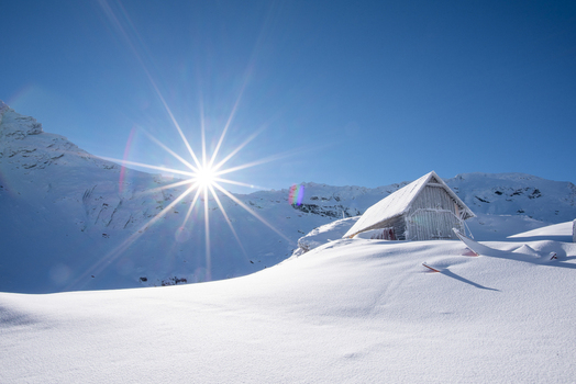 View of a small wooden cabin nestled in the snow covered mountains of the Transfagarasan pass under a bright sunburst in the clear blue sky Curtea de Arges Judetul Arges Romania. View of a small wooden cabin nestled in the snow covered mountains of the Transfagarasan pass under a bright sunburst in the clear blue sky Curtea de Arges Judetul Arges Romania.