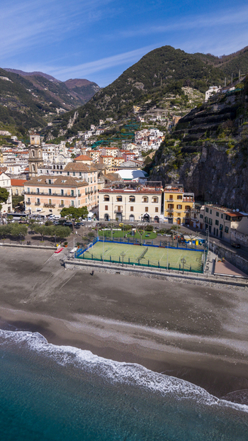 Aerial view of sun kissed buildings cascading down to a dark sandy beach meeting the turquoise sea nestled beneath verdant mountains Minori Campania Italy. Aerial view of sun kissed buildings cascading down to a dark sandy beach meeting the turquoise sea nestled beneath verdant mountains Minori Campania Italy.
