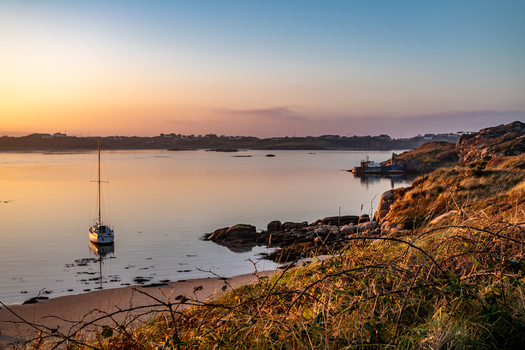 View of a sailboat anchored in the calm waters of the bay at sunset with rocky shores and grassy foreground in Gortnasade County Donegal Ireland. View of a sailboat anchored in the calm waters of the bay at sunset with rocky shores and grassy foreground in Gortnasade County Donegal Ireland.