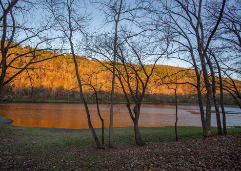 View of golden light sweeps across the water and hillside framed by the stark silhouettes of bare trees on the grassy bank Fort Smith Arkansas United States. View of golden light sweeps across the water and hillside framed by the stark silhouettes of bare trees on the grassy bank Fort Smith Arkansas United States.