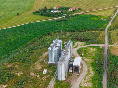 Aerial view of metallic grain silos situated in a rural landscape with surrounding green fields and dirt roads in Poland.