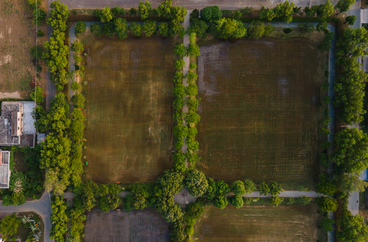 Aerial view of two large rectangular grass fields separated by a tree lined path in a park with surrounding greenery and urban structures in Lahore Punjab Pakistan.