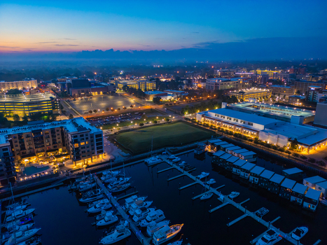 Aerial view of Riverfront Park at dusk featuring a marina filled with boats illuminated modern buildings and city lights reflecting on the water in Wilmington North Carolina United States.