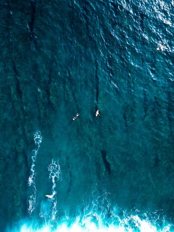 Aerial view of surfers on surfboards in the deep blue ocean waters of Pantai Nunggalan with white sea foam and clear waves Kecamatan Kuta Selatan Bali Indonesia.