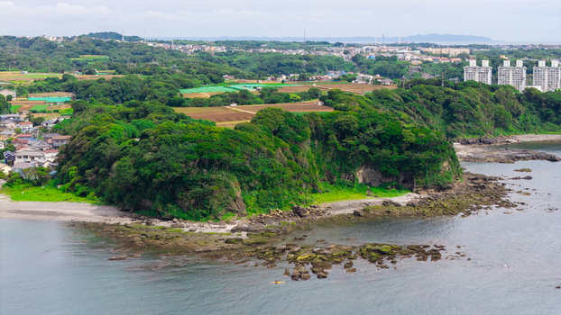 Aerial view of a lush green forested headland extending into the sea with rocky outcrops a sandy beach and a small kayak paddling in the water near the coast of Japan. Aerial view of a lush green forested headland extending into the sea with rocky outcrops a sandy beach and a small kayak paddling in the water near the coast of Japan.