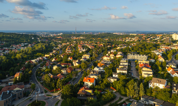 Aerial view of residential neighborhoods with red tiled roofs winding roads lush green trees and a distant industrial skyline in Gdansk Pomeranian Voivodeship Poland. Aerial view of residential neighborhoods with red tiled roofs winding roads lush green trees and a distant industrial skyline in Gdansk Pomeranian Voivodeship Poland.