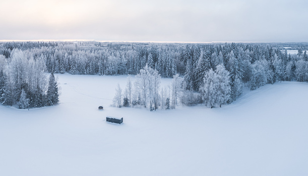 Aerial view of a small dark cabin in a vast snow covered field surrounded by frost covered trees and a dense forest in Latvia. Aerial view of a small dark cabin in a vast snow covered field surrounded by frost covered trees and a dense forest in Latvia.