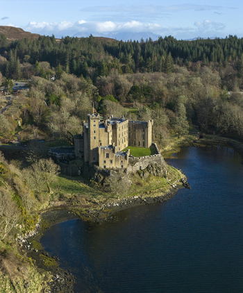Aerial view of Dunvegan Castle and Gardens situated on a rocky outcrop overlooking the blue waters of Loch Dunvegan surrounded by lush forests in Isle of Skye Scotland United Kingdom. Aerial view of Dunvegan Castle and Gardens situated on a rocky outcrop overlooking the blue waters of Loch Dunvegan surrounded by lush forests in Isle of Skye Scotland United Kingdom.