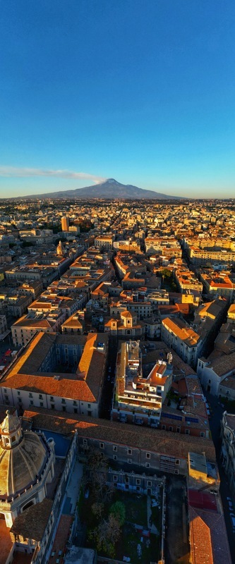 Aerial view of the historic city center with dense rooftops and the majestic Mount Etna volcano under a clear blue sky at sunset in Catania Sicily Italy. Aerial view of the historic city center with dense rooftops and the majestic Mount Etna volcano under a clear blue sky at sunset in Catania Sicily Italy.