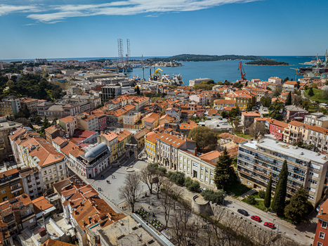 Aerial view of terracotta rooftops cascading towards the shimmering Adriatic Sea near the port where cranes stand tall against the horizon Pula Istria County Croatia.