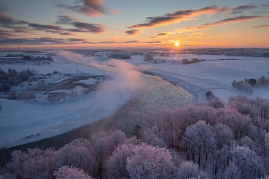 Aerial view of the Lielupe river flowing through a snow covered landscape with frost covered trees and mist rising under a golden sunrise in Bauska Ziedoni Latvia.