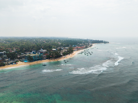 Aerial view of the sandy Hikkaduwa Beach with turquoise waters breaking waves and beachfront buildings surrounded by palm trees in Hikkaduwa Southern Province Sri Lanka. Aerial view of the sandy Hikkaduwa Beach with turquoise waters breaking waves and beachfront buildings surrounded by palm trees in Hikkaduwa Southern Province Sri Lanka.