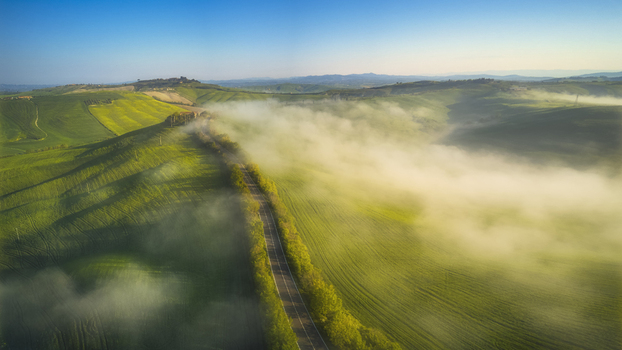Aerial view of rolling green hills and a winding road shrouded in morning mist under a clear blue sky in Tuscany Italy.