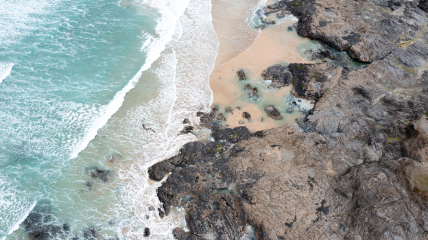 Aerial view of Constantine Bay with turquoise waves crashing against dark jagged rocks and a sandy shoreline Padstow Cornwall United Kingdom. Aerial view of Constantine Bay with turquoise waves crashing against dark jagged rocks and a sandy shoreline Padstow Cornwall United Kingdom.