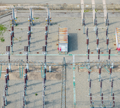 Aerial view of high voltage switchgear and electrical substation infrastructure at a power generation facility with metal structures and gravel ground in Italy.