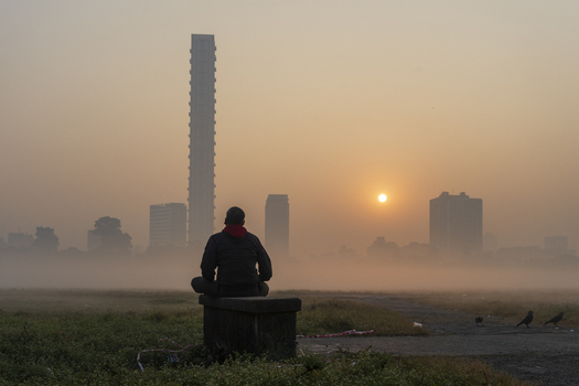 View of a solitary figure sits in serene contemplation as the sun rises over a misty cityscape with towering buildings Kolkata West Bengal India. View of a solitary figure sits in serene contemplation as the sun rises over a misty cityscape with towering buildings Kolkata West Bengal India.