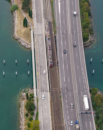 Aerial view of the Melide Causeway with a train traveling on tracks alongside a highway with cars surrounded by the waters of Lake Lugano in Melide Ticino Switzerland. Aerial view of the Melide Causeway with a train traveling on tracks alongside a highway with cars surrounded by the waters of Lake Lugano in Melide Ticino Switzerland.