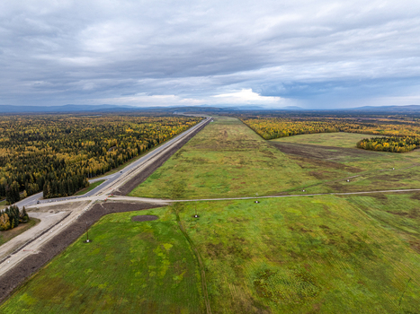 Aerial view of a long highway cutting through a vast landscape of autumn colored forests and green fields under a cloudy sky in Moose Creek Alaska United States.