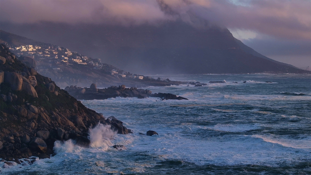 Aerial view of the rocky coastline with crashing waves and houses on a hillside under misty mountains in Cape Town Western Cape South Africa. Aerial view of the rocky coastline with crashing waves and houses on a hillside under misty mountains in Cape Town Western Cape South Africa.