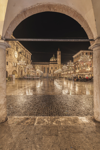 View of the illuminated Piazza del Popolo at night with historic buildings and reflections on the wet cobblestone square in Ascoli Piceno Marche Italy.