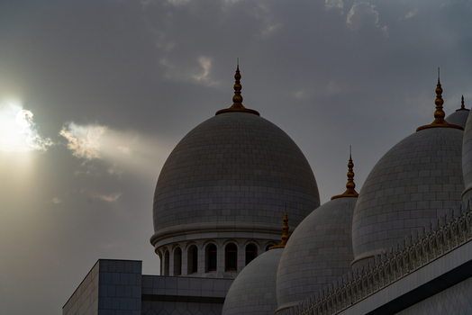 View of the white marble domes of a grand mosque with gold finials under a dramatic cloudy sky with sun rays in Abu Dhabi Abu Dhabi United Arab Emirates. View of the white marble domes of a grand mosque with gold finials under a dramatic cloudy sky with sun rays in Abu Dhabi Abu Dhabi United Arab Emirates.