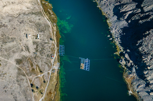 Aerial view of Blind Lake Sarfaranga Shigar with fish farming cages in clear turquoise water surrounded by arid rocky terrain and dirt paths in Skardu Gilgit Pakistan.