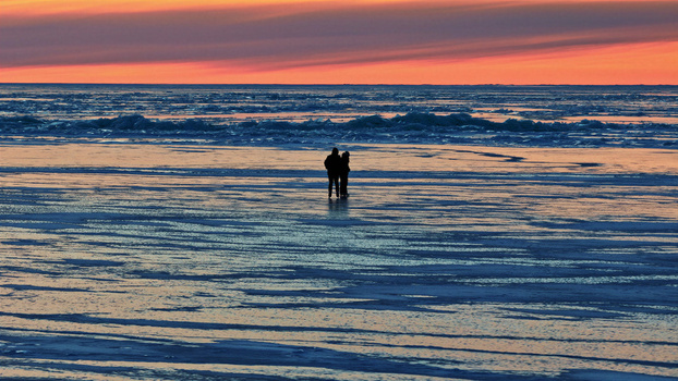 Aerial view of two people standing on a vast frozen sea surface under a vibrant orange and blue sunset sky in Latvija. Aerial view of two people standing on a vast frozen sea surface under a vibrant orange and blue sunset sky in Latvija.