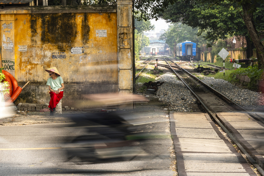 Hanoi Vietnam 11 November 2025 View of a woman in a conical hat sitting by a yellow wall near the Hanoi Train Street with railway tracks and a passing train.