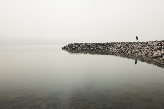 Saxby Estonia 01 June 2025 View of a person standing on the rocky Saxby Coast breakwater extending into the calm still waters under a soft overcast sky.