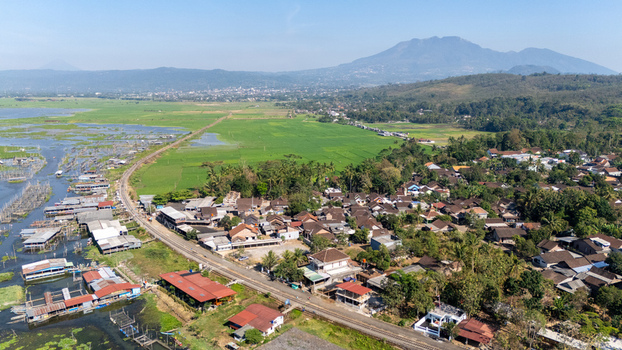 Aerial view of Lake Rawa Pening featuring a village green rice fields and floating structures along the water with mountains in Waduk Rawapening Central Java Indonesia. Aerial view of Lake Rawa Pening featuring a village green rice fields and floating structures along the water with mountains in Waduk Rawapening Central Java Indonesia.