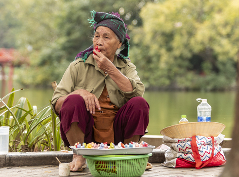 Hanoi Ha Noi Vietnam 11 November 2025 View of an elderly woman sitting by a lake painting small colorful figurines with a basket of crafts and a conical hat nearby.