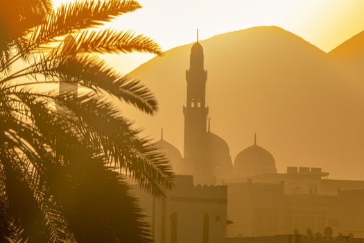 View of a mosque silhouette with minarets and domes at sunset framed by palm tree leaves in Muscat Muscat Governorate Oman. View of a mosque silhouette with minarets and domes at sunset framed by palm tree leaves in Muscat Muscat Governorate Oman.